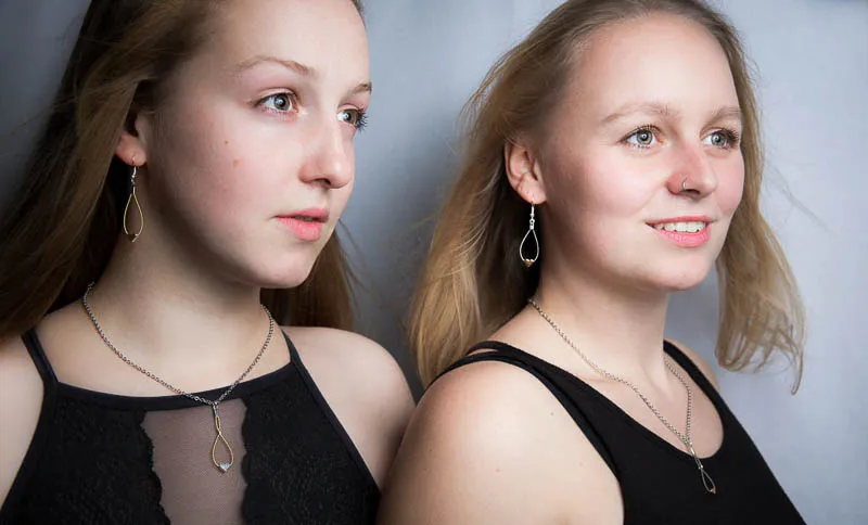 Fashion Portrait mit zwei Frauen. Die Frau links im Bild hat brünettes mittellanges Haar, die Frau rechts im Bild hat blondes mittellanges Haar. Beide Frauen blicken an der Kamera vorbei. Sie tragen eleganten Schmuck mit kleinen Herzen. Ihre Haare wehen leicht im Wind. Beide Frauen tragen ärmellose schwarze Shirts.